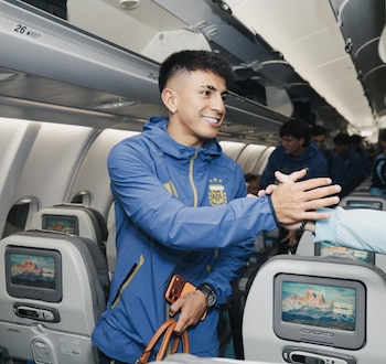 Un futbolista joven con un chándal azul de la Selección Argentina sonríe y choca las manos dentro de un avión, con otros pasajeros al fondo