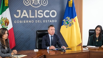 Pablo Lemus sentado ante un micrófono en una mesa de madera, flanqueado por dos mujeres y banderas oficiales