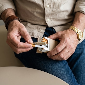 Primer plano de manos de una persona sacando un cigarrillo de tabaco y un porro de cannabis de una cajetilla abierta. Lleva una pulsera de cuero y reloj dorado.