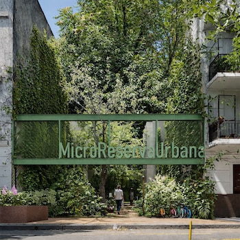 Entrada a la MicroReserva Urbana con cartel verde, abundante vegetación, una persona caminando y bicicletas estacionadas. Edificios cubiertos de plantas a los lados