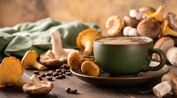 Una taza de café verde con platillo en una mesa de madera, rodeada de hongos shiitake, rebozuelos y otros, granos de café y una tela verde al fondo.