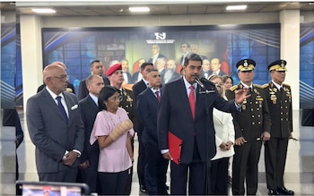 Un hombre en traje habla al micrófono, flanqueado por personas en trajes y uniformes militares, y una mujer con el brazo vendado. Al fondo, un mural histórico