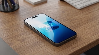 Un smartphone gris está colocado sobre un escritorio de madera. La pantalla muestra un fondo abstracto en blanco, celeste y azul con el número '27' en el centro.