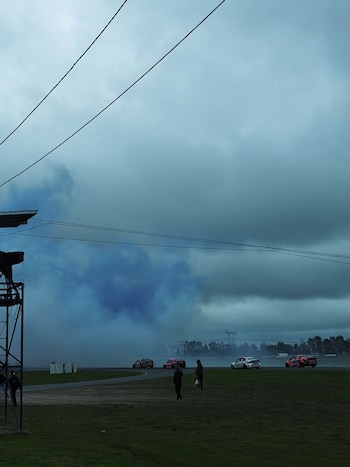 En un circuito, mujeres y niños con auriculares disfrutan del evento. Se ve un coche de carreras azul y una mujer con abrigo rojo en el pit lane