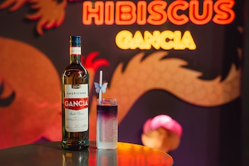 Botella de aperitivo Gancia y vaso con bebida de dos tonos y pajita con mariposa sobre una mesa frente a un cartel de neón