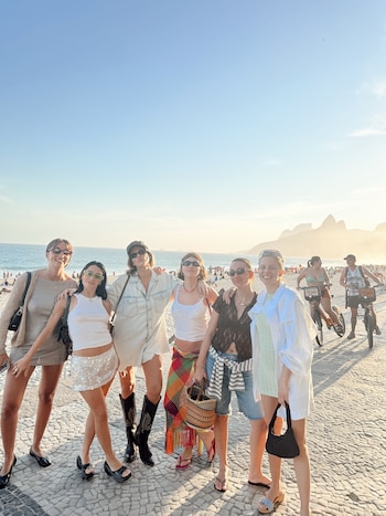 Seis mujeres posan en un paseo peatonal junto a la playa con gafas de sol y ropa de verano, con el mar y montañas al fondo