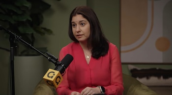 Una mujer de cabello oscuro y camisa rosa está sentada, hablando frente a un micrófono con un logo amarillo en un estudio con fondo verde