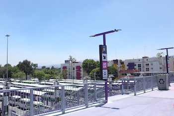Vista de un moderno paradero de autobuses con barandales morados y grises, un poste con señalización "ANDÉN A" y camionetas, bajo cielo azul