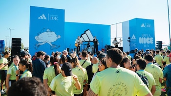 Multitud frente a escenario azul con banda tocando y banners de Adidas Supernova 10K. Muchas personas visten camisetas amarillas, bajo cielo despejado
