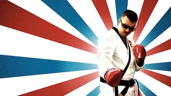 Hombre con bigote, gafas de sol y uniforme blanco de taekwondo con cinturón negro y guantes de boxeo rojos frente a un fondo de rayas radiales rojas, blancas y azules