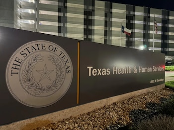 Señal de Texas Health & Human Services y El Estado de Texas, frente a un edificio moderno. Incluye las banderas de Texas y de Estados Unidos, y rocas en el suelo