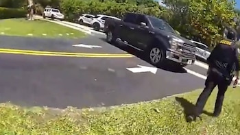 Un oficial de policía con uniforme oscuro camina por el césped. A su izquierda, una carretera con líneas amarillas, un camión oscuro y coches de policía