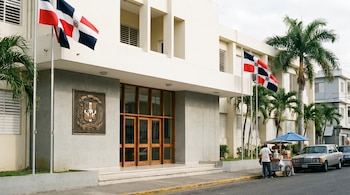 Entrada de edificio escolar blanco con banderas dominicanas, escudo nacional, palmeras, un vendedor ambulante con carrito y un coche plateado estacionado.