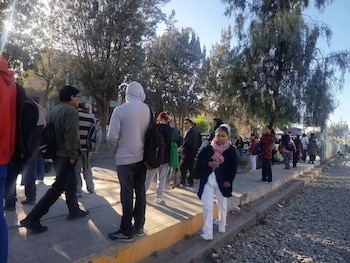 Grupo de personas en una acera y una calle de grava, rodeados de árboles, con sol brillante en el fondo y sombras proyectadas