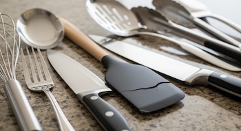 Primer plano de varios utensilios de cocina metálicos como batidor, tenedor, cuchillos y espátulas, incluyendo una de goma agrietada, sobre una mesada granulada.