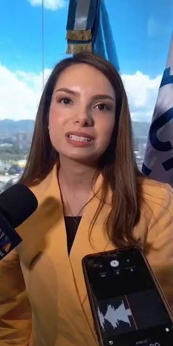 Una mujer con chaqueta amarilla habla ante un micrófono junto a un teléfono inteligente que exhibe una onda de audio en pantalla