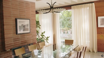 Comedor moderno con mesa de cristal, sillas de madera, pared de ladrillo, cuadro, planta y grandes ventanales con cortinas blancas translúcidas