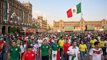 Multitud de personas con camisetas de fútbol y banderas camina por una plaza en Ciudad de México, con edificios históricos decorados y una gran bandera mexicana.