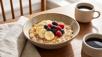 Tazón de avena cortada decorado con rodajas de plátano, frambuesas y arándanos, junto a dos tazas con café negro y una servilleta de tela.