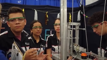 Cuatro jóvenes con gafas de seguridad y uniformes oscuros observan un robot con estructuras metálicas, cables y una luz naranja. Hay decoraciones colgantes de fondo