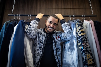 Hombre con barba, sonriente, posando detrás de un perchero con ropa, sosteniendo la barra con ambas manos. Fondo de pared de madera