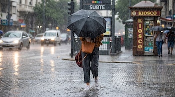 Vista a plano corto de una persona con paraguas negro caminando por una calle de adoquines empapada por la lluvia, con charcos y salpicaduras.