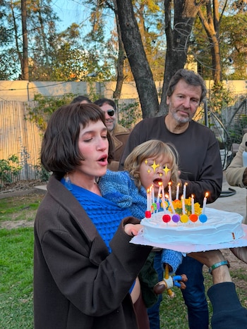 Cande Vetrano sostiene a un niño que observa una torta de cumpleaños con velas encendidas, con adultos y árboles de fondo en un jardín