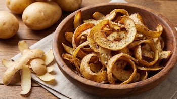 Un cuenco de madera lleno de chips de cáscara de papa con especias y sal, junto a papas enteras y cáscaras peladas sobre una tela y madera.