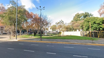 Imagen de Parque España en Barracas, mostrando una zona verde con árboles, una farola y una estatua cercada, junto a una calle asfaltada y edificios