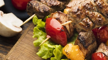Primer plano de brochetas de carne y pimientos asados (rojos y amarillos) sobre lechuga fresca en una tabla de madera, con un champiñón al fondo