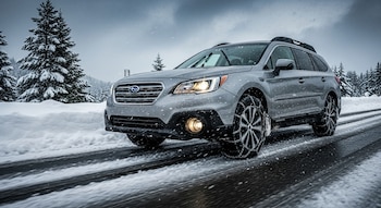 Un automóvil Subaru gris con cadenas de nieve en las ruedas avanza por una carretera nevada, bajo una nevada, con árboles cubiertos de nieve.