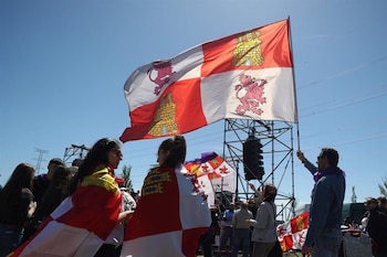 Una gran bandera de Castilla y León ondea en el aire sobre una multitud de personas, algunas con banderas más pequeñas, en un día soleado y claro al aire libre