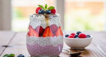 Primer plano de un frasco de pudín de chía en capas con fresas, frambuesas, arándanos y hojas de menta en una mesa de madera.