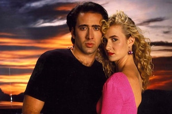 Hombre de cabello oscuro con camiseta negra y mujer rubia de cabello rizado con blusa rosa posan contra un atardecer anaranjado y morado