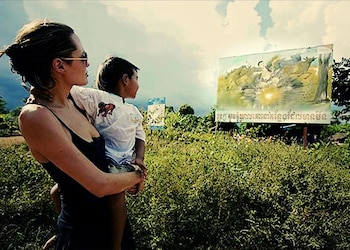 Una mujer con gafas de sol sostiene a un niño en brazos frente a un cartel ilustrado con texto en jemer en un campo con vegetación