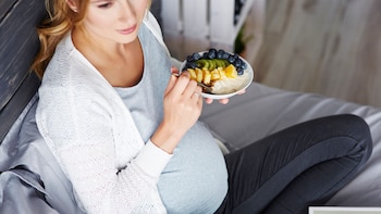 Mujer embarazada sentada, vestida con suéter blanco y blusa gris, sosteniendo un tazón blanco con arándanos, kiwi, naranja y yogur, comiendo con una cuchara