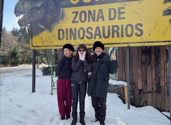 Tres personas sonrientes en la nieve frente a un cartel amarillo grande con un dinosaurio y el texto ZONA DE DINOSAURIOS. Hay árboles y una carretera detrás