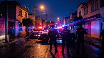 Siluetas de tres agentes de espaldas frente a patrullas con luces rojas y azules en una calle mojada de noche.