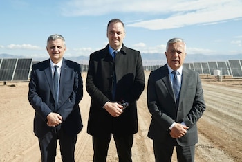 Três homens de terno e gravata estão diante de um campo de painéis solares sob um céu azul claro. A terra está seca e montanhas podem ser vistas ao fundo.