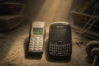 Dos teléfonos móviles antiguos, un Nokia y un BlackBerry, cubiertos de polvo sobre una mesa iluminada por un haz de luz.