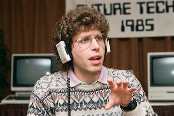 Hombre con anteojos, suéter y auriculares de diadema hace un gesto frente a dos monitores antiguos y un cartel de Future Tech 1985