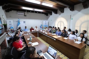 Sala de reuniones con personas en una mesa en U, ordenador portátil, calculadora, vasos, papeles, bandera de Guatemala, pancartas de MAGA y DICORER