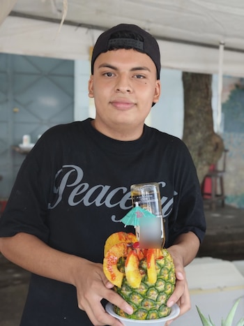 Hombre y mujer asiáticos sonriendo en primer plano, la mujer sostiene un letrero con forma de piña que dice "Feria de la Piña 2026 Santa María Ostuma"