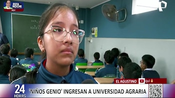 Una estudiante con gafas mira al frente, sentada en un aula. Hay otros estudiantes en pupitres, una pizarra y un ventilador
