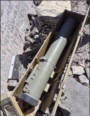 Un misil militar grande, de color verde oliva, yace en una caja de madera rota. Está rodeado de escombros de hormigón gris y rocas en un área destruida