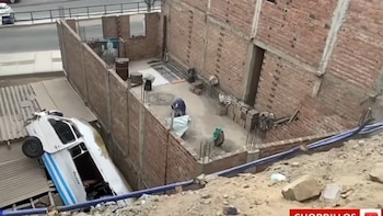 Una combi blanca y azul incrustada de lado entre el techo de una vivienda y una pared de ladrillos en una ladera de tierra