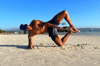 Hombre practicando yoga en un tapete azul junto a un río verde. Viste camiseta beige y shorts azules. Al fondo, edificios coloridos y montañas