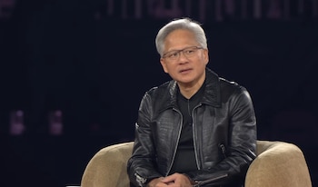 Hombre con cabello gris, gafas, chaqueta de cuero negra y camisa oscura, sentado en un sillón de tela clara, con fondo oscuro