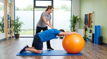 Hombre de rodillas sobre colchoneta azul, brazos estirados sobre un balón naranja. Una fisioterapeuta de pie detrás, con las manos en su espalda. Sala con grandes ventanas.