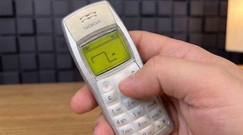 Una mano sostiene un teléfono móvil Nokia plateado con pantalla verde que muestra el juego Snake y un teclado numérico.